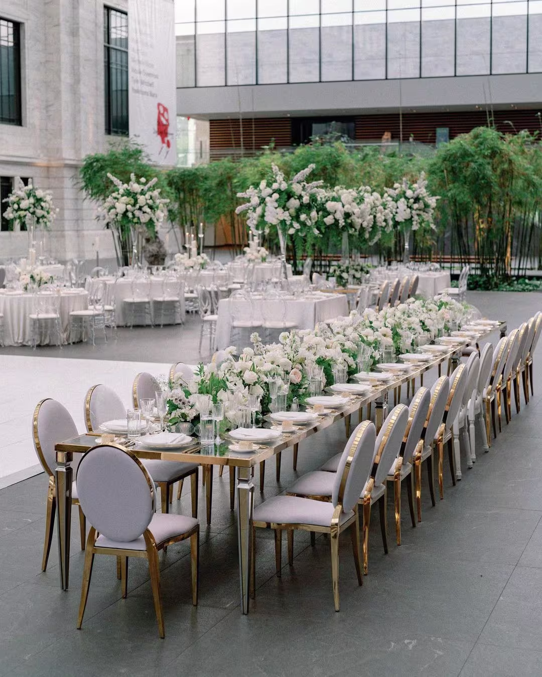 Outdoor elegant wedding dining setup with long table, white chairs, and floral arrangements in a modern venue